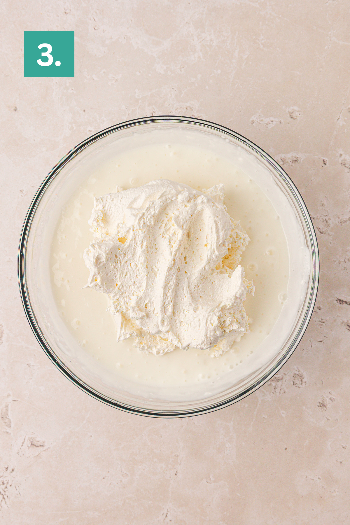 A glass bowl filled with whipped cream sitting on top of a white creamy liquid, placed on a light beige marble surface. A green box with the number 3 appears in the top left corner.