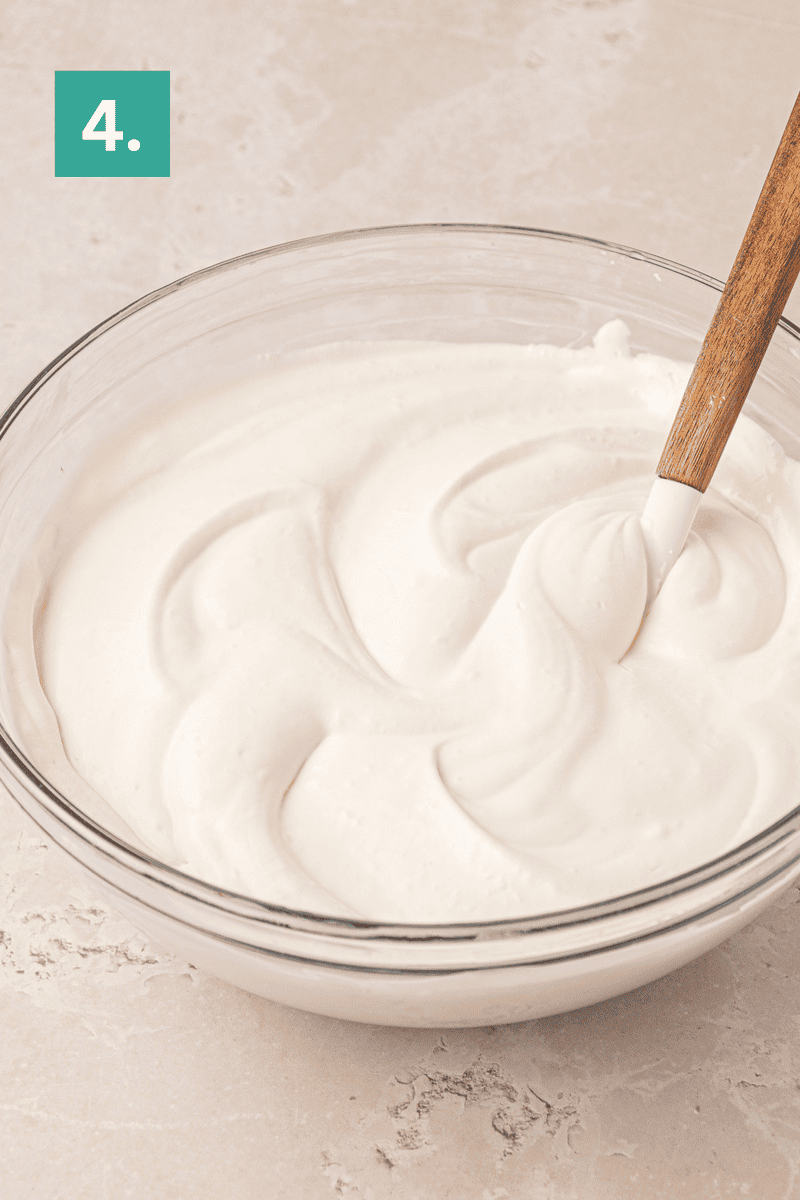 A glass bowl filled with creamy, whipped mixture sits on a light surface. A spatula with a wooden handle rests in the bowl. A green box with the number 4 is in the top left corner.