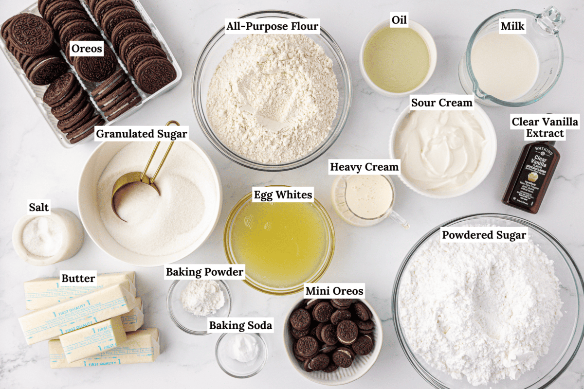 A top-down view of labeled baking ingredients on a white surface, including flour, sugar, butter, egg whites, milk, oil, heavy cream, sour cream, powdered sugar, Oreos, mini Oreos, salt, baking powder, baking soda, and vanilla extract.