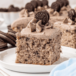 A square slice of cookies and cream cake topped with cookies and cream frosting and a mini Oreo cookie, served on a white plate with more cake and cookies visible in the background.