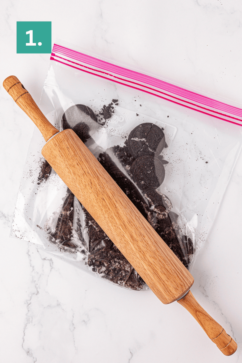 A wooden rolling pin is resting on a ziplock bag filled with crushed chocolate sandwich cookies on a white surface. A green box with the number 1 is in the top left corner.