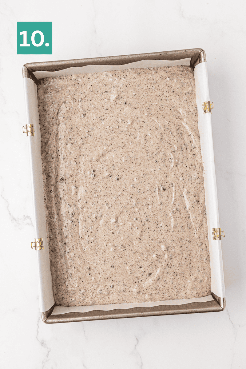 A parchment-lined square baking pan filled with speckled batter, ready to be baked. The pan is secured with metal clips, and a green square with the number 10 is in the top left corner.