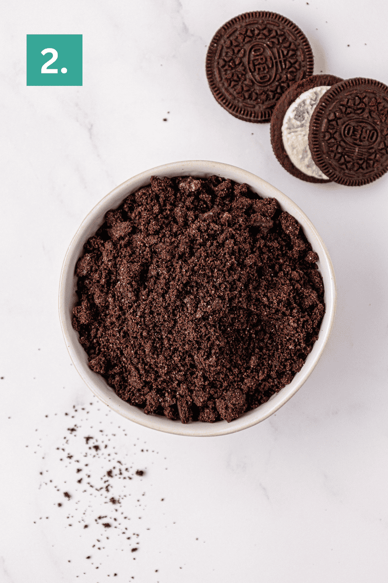 A bowl filled with finely crushed chocolate sandwich cookies sits on a white surface, with a few whole and partially split cookies and crumbs scattered nearby. A green square in the corner displays the number 2.