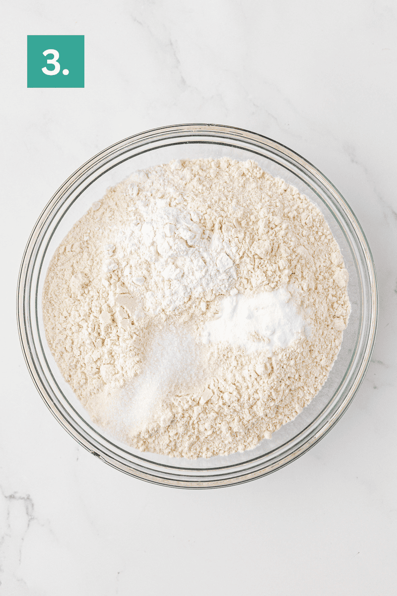 A glass bowl filled with flour, sugar, baking powder, and baking soda sits on a white marble surface. A green box in the top left corner displays the number 3.