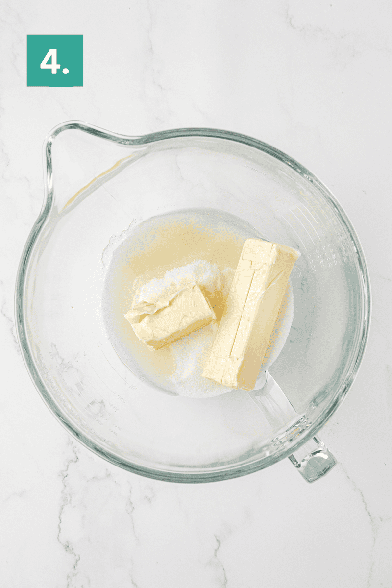 A glass mixing bowl containing two sticks of butter, sugar, and some liquid on a white marble surface. A green box with the number 4 appears in the upper left corner.
