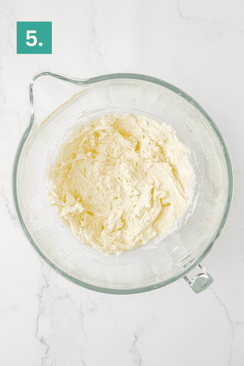 A glass mixing bowl filled with creamed butter and sugar sits on a white marble surface. A green box labeled “5.” is in the top left corner, indicating step five of a recipe.