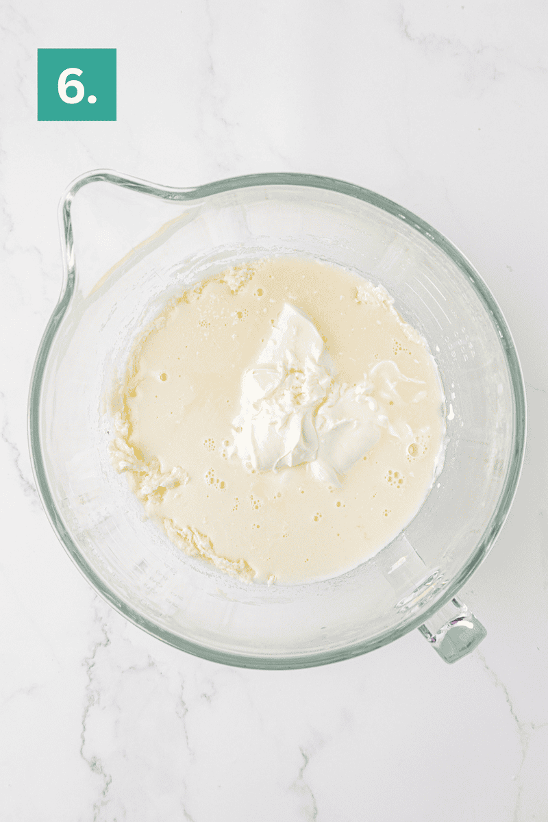 A glass mixing bowl with wet and dry batter ingredients, including a dollop of sour cream, on a white marble surface. The top left corner has a green box with the number 6.