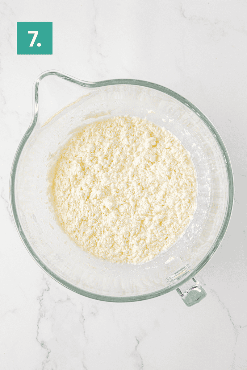 A glass mixing bowl filled with a crumbly, pale mixture sits on a white marble surface. A green box labeled 7. is in the top left corner of the image.