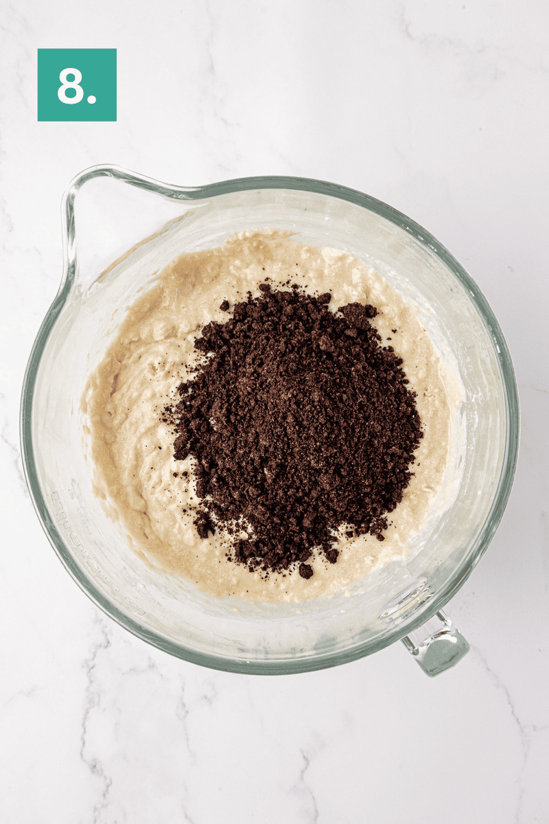 A glass mixing bowl with cake batter and a pile of crushed chocolate cookies on top is shown on a white surface. A green box with the number 8 is in the top left corner.