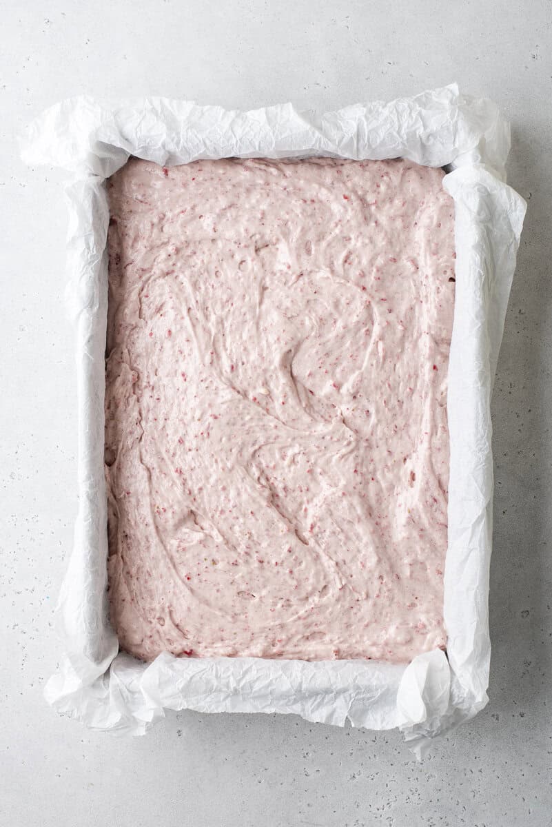 A rectangular baking pan lined with white parchment paper, filled with an uncooked, light pink, textured batter spread evenly to the edges.