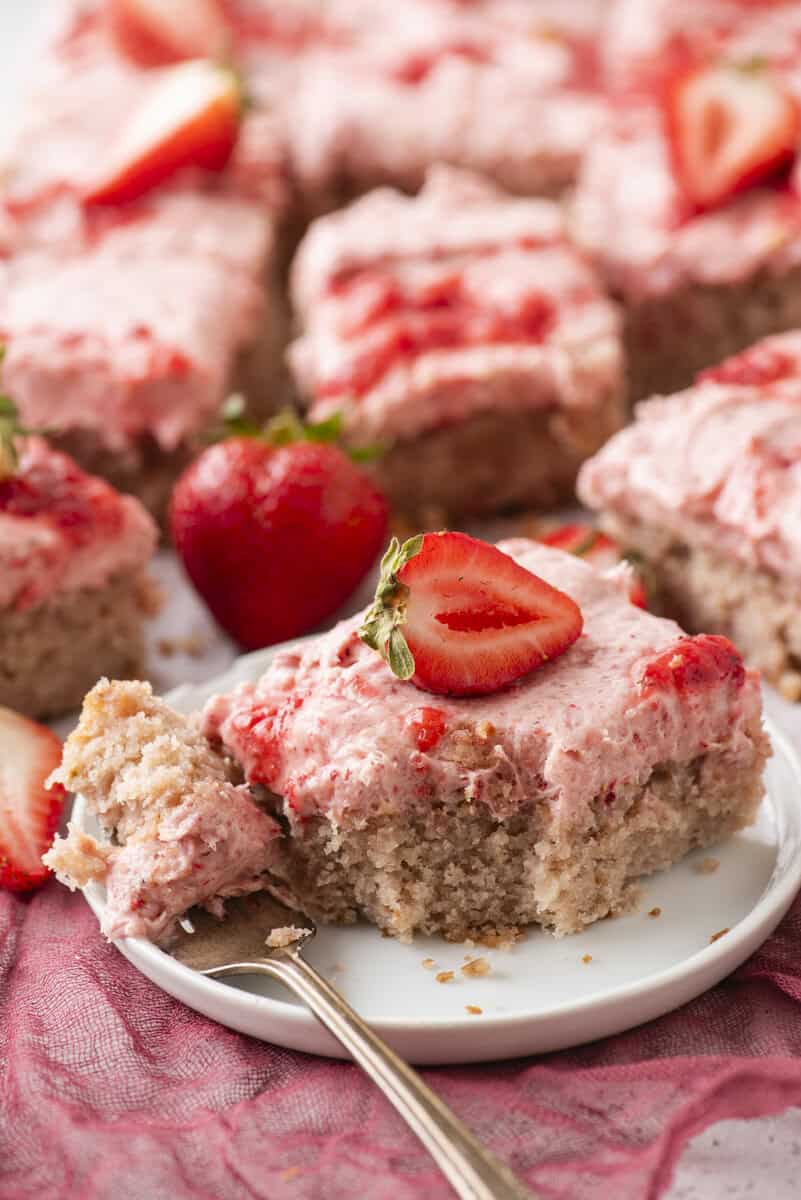 A square piece of strawberry cake with pink frosting and a fresh strawberry slice on top sits on a white plate. A fork has taken a bite from the cake, and more frosted cake pieces are in the background.