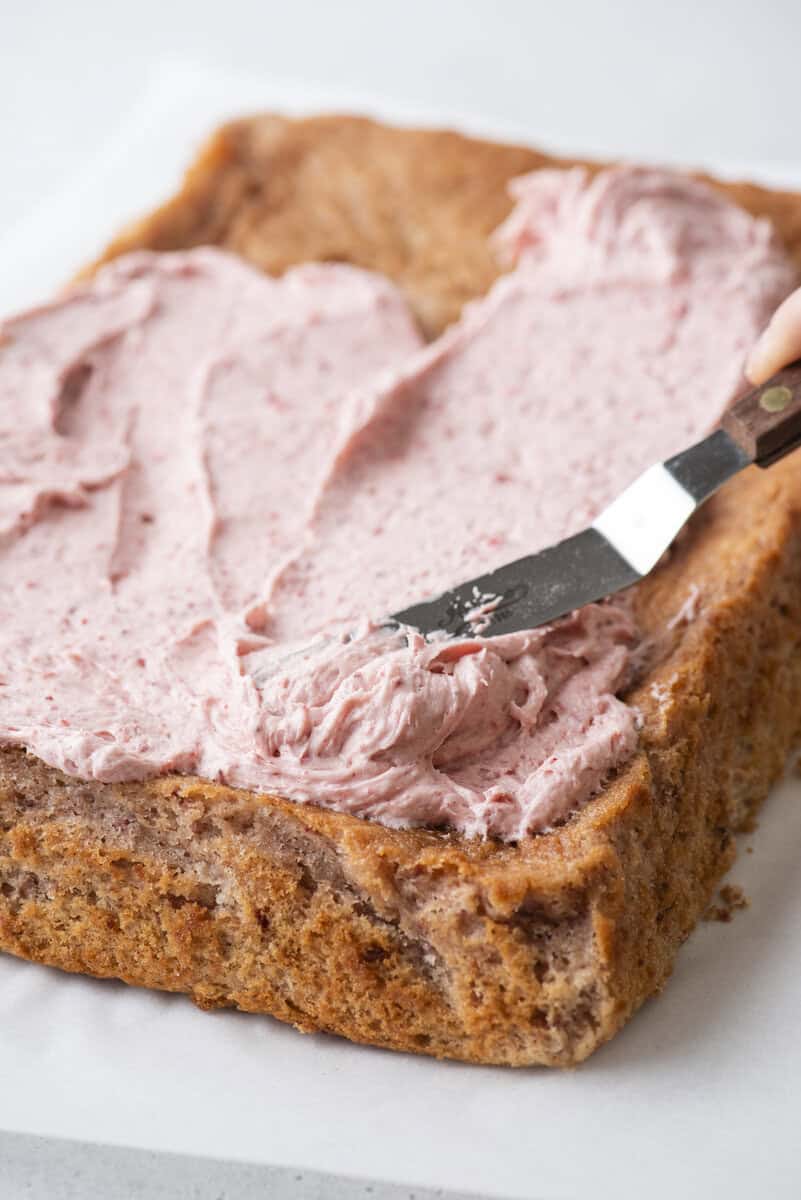 A hand uses an offset spatula to spread pink frosting over a rectangular, rustic-looking cake on a white surface.