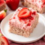 A square slice of strawberry cake with pink strawberry frosting and a halved fresh strawberry on top, served on a white plate with whole and cut strawberries in the background.
