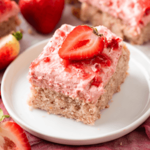 A square slice of strawberry cake with pink strawberry frosting and a halved fresh strawberry on top, served on a white plate with whole and cut strawberries in the background.