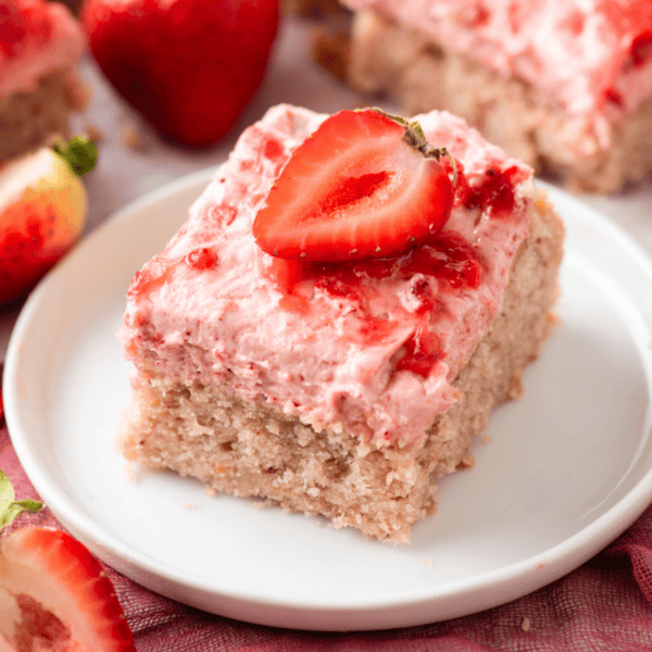 A square slice of strawberry cake with pink strawberry frosting and a halved fresh strawberry on top, served on a white plate with whole and cut strawberries in the background.