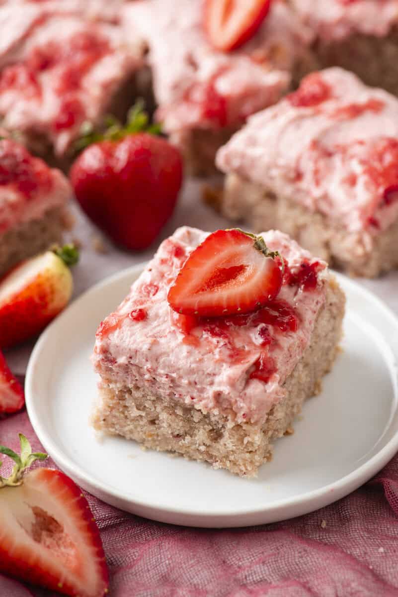 A square piece of strawberry cake with pink strawberry frosting and a halved fresh strawberry on top sits on a white plate. Whole and sliced strawberries and more cake pieces are in the background.