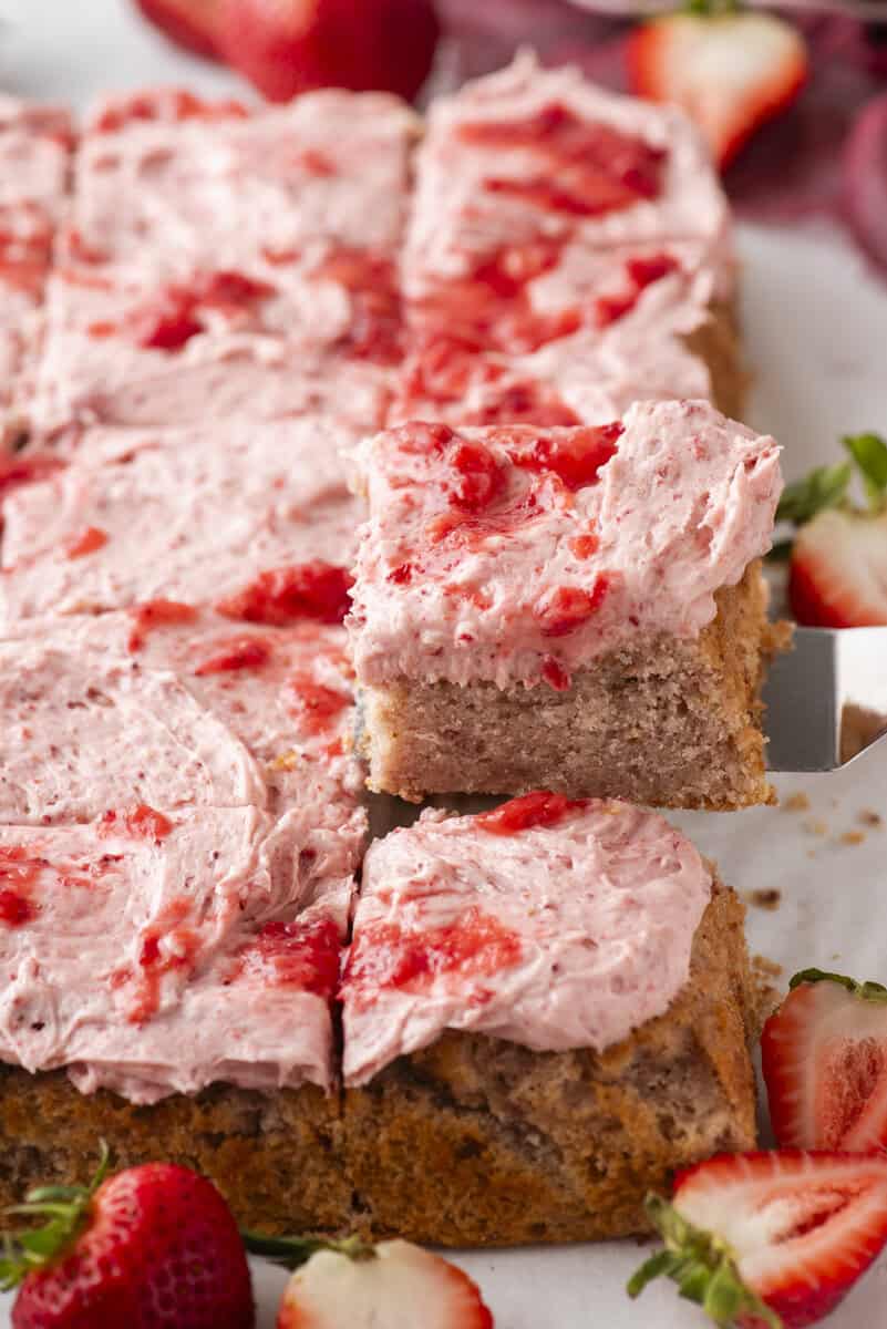 A close-up of a sheet cake cut into squares with pink strawberry frosting and fresh strawberry pieces on top. One slice is lifted, revealing the moist, airy cake. Whole and sliced strawberries are scattered around.