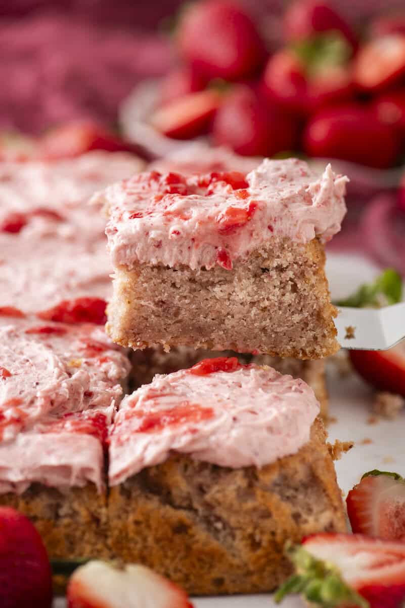 A close-up of a square slice of strawberry cake with pink strawberry frosting being lifted from a tray. Fresh strawberries are visible in the background and around the cake.