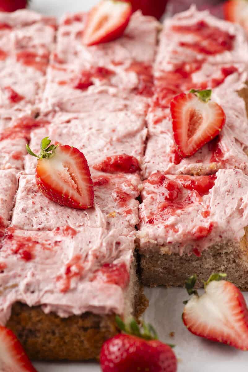 A close-up of sliced sheet cake topped with pink strawberry frosting and fresh strawberry halves, with visible cake texture and strawberries scattered around.