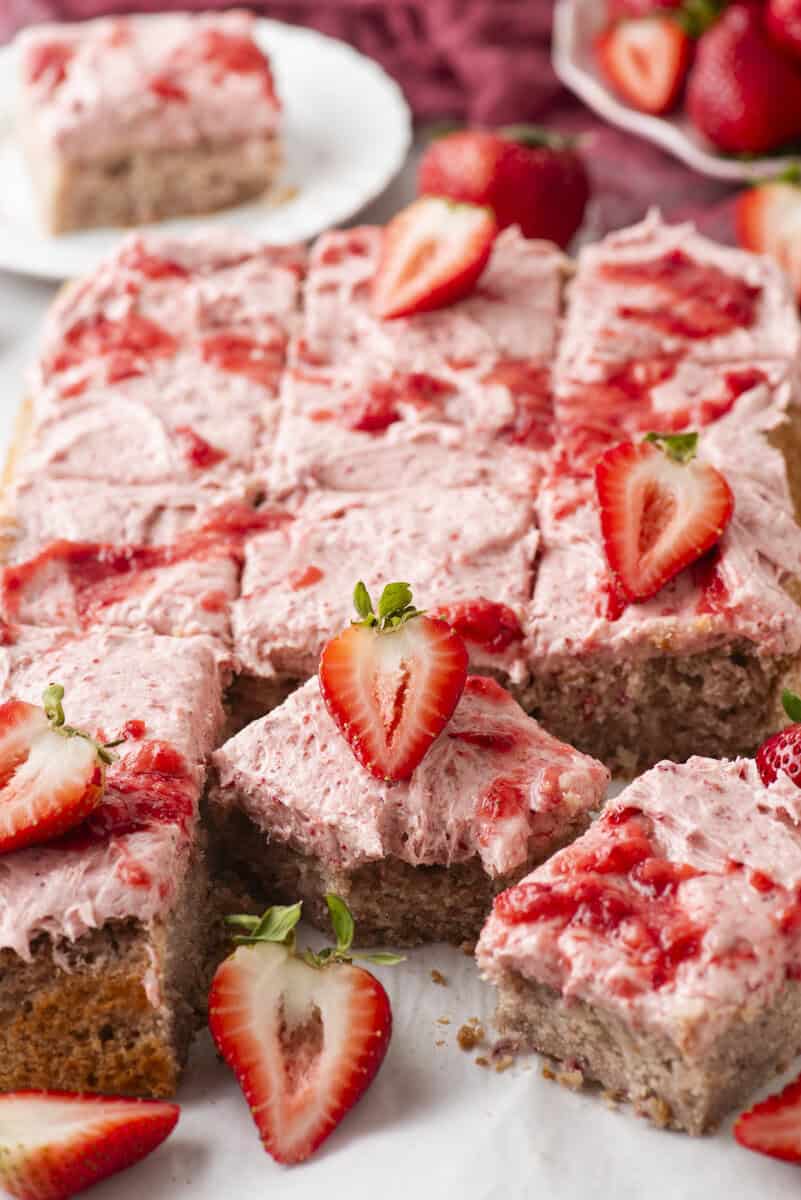 A sheet cake topped with pink strawberry frosting and fresh strawberry slices, cut into squares, with one piece lifted up. Whole and sliced strawberries are scattered around the cake.