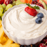 A white bowl filled with creamy fruit dip, topped with a strawberry and two blueberries, surrounded by fresh fruit pieces including pineapple, grapes, berries, and melon.