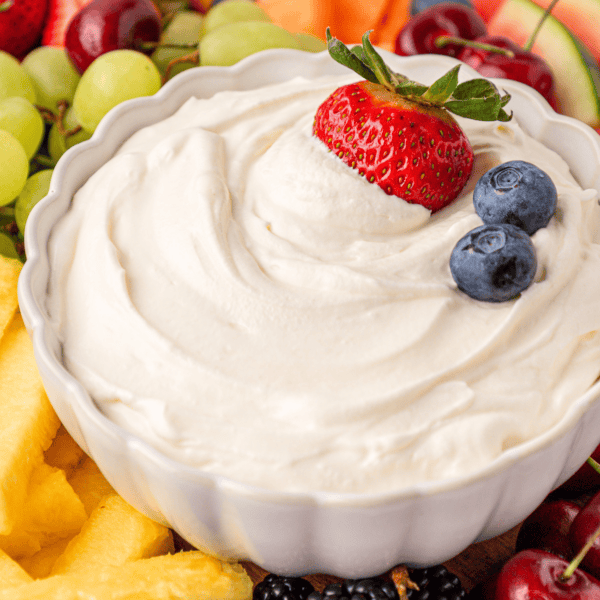 A white bowl filled with creamy fruit dip, topped with a strawberry and two blueberries, surrounded by fresh fruit pieces including pineapple, grapes, berries, and melon.