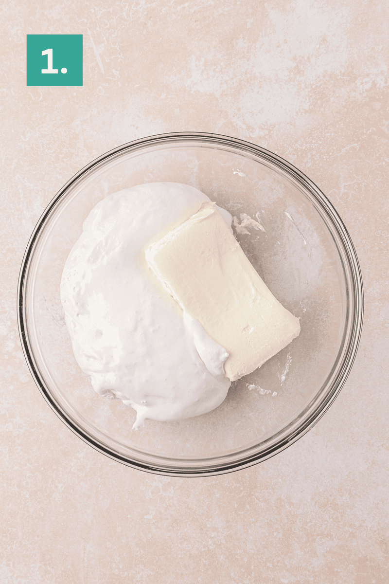 A glass bowl containing a block of cream cheese and a large dollop of marshmallow fluff, on a light countertop. A green box with the number 1 is in the top left corner.