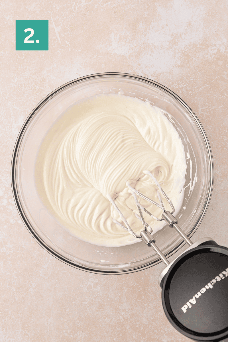 A glass bowl filled with smooth, whipped cream cheese mixture is being mixed with an electric hand mixer. The background is a light countertop, and a green box with the number 2 is in the top left corner.