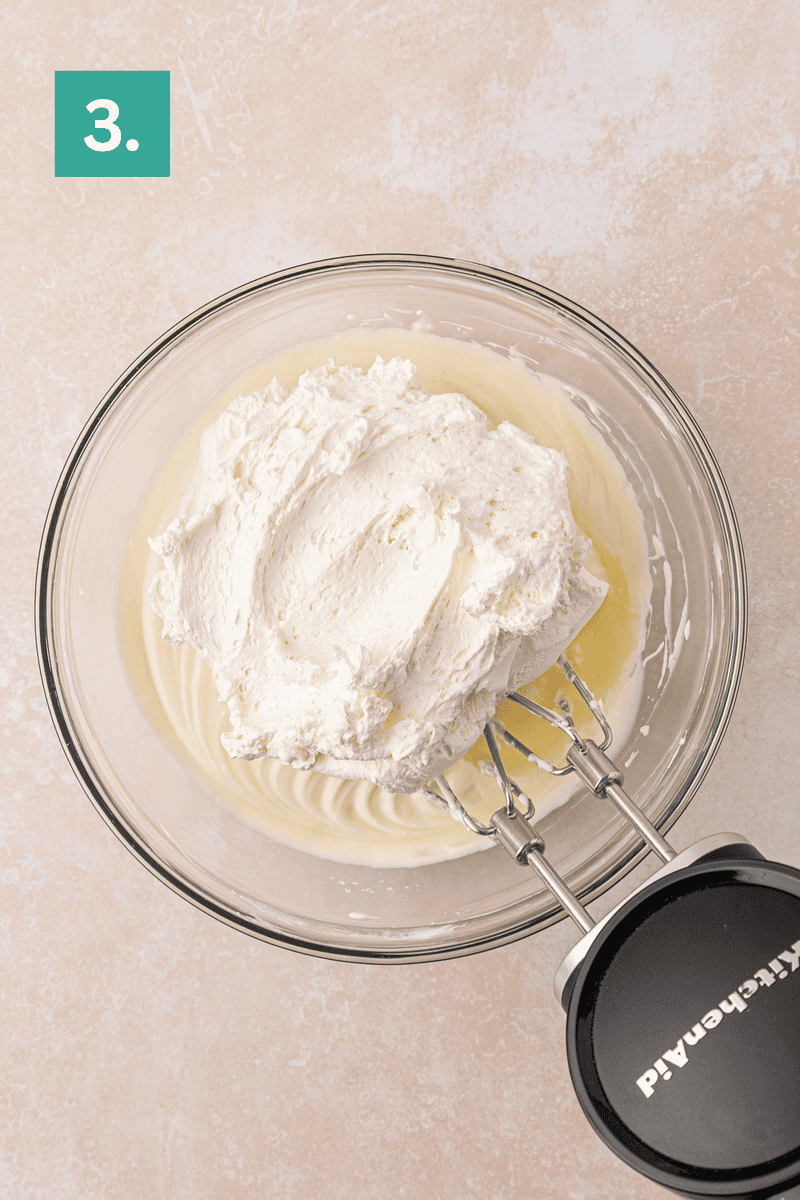 A glass bowl with whipped cream being mixed, seen from above. An electric mixer is in the bowl, and a green box with the number 3 is in the top left corner.