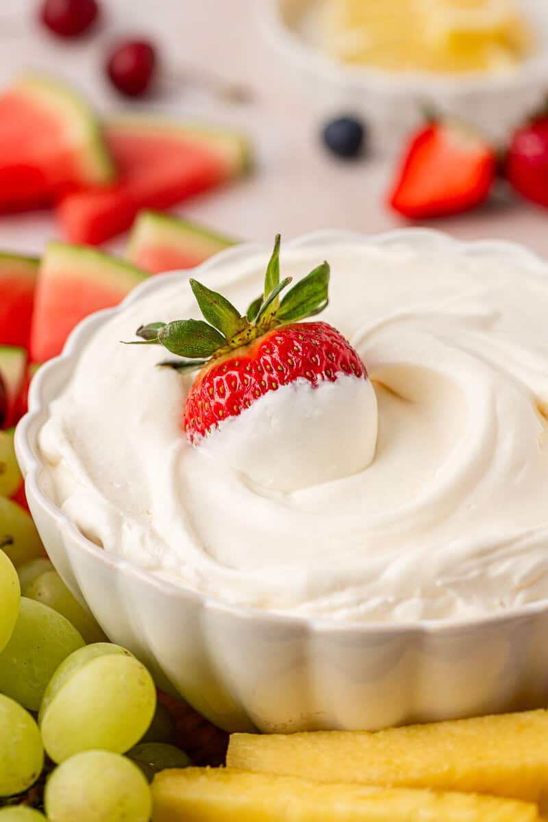 A white bowl filled with creamy fruit dip, topped with a whole strawberry, surrounded by fresh grapes, pineapple, and watermelon slices.