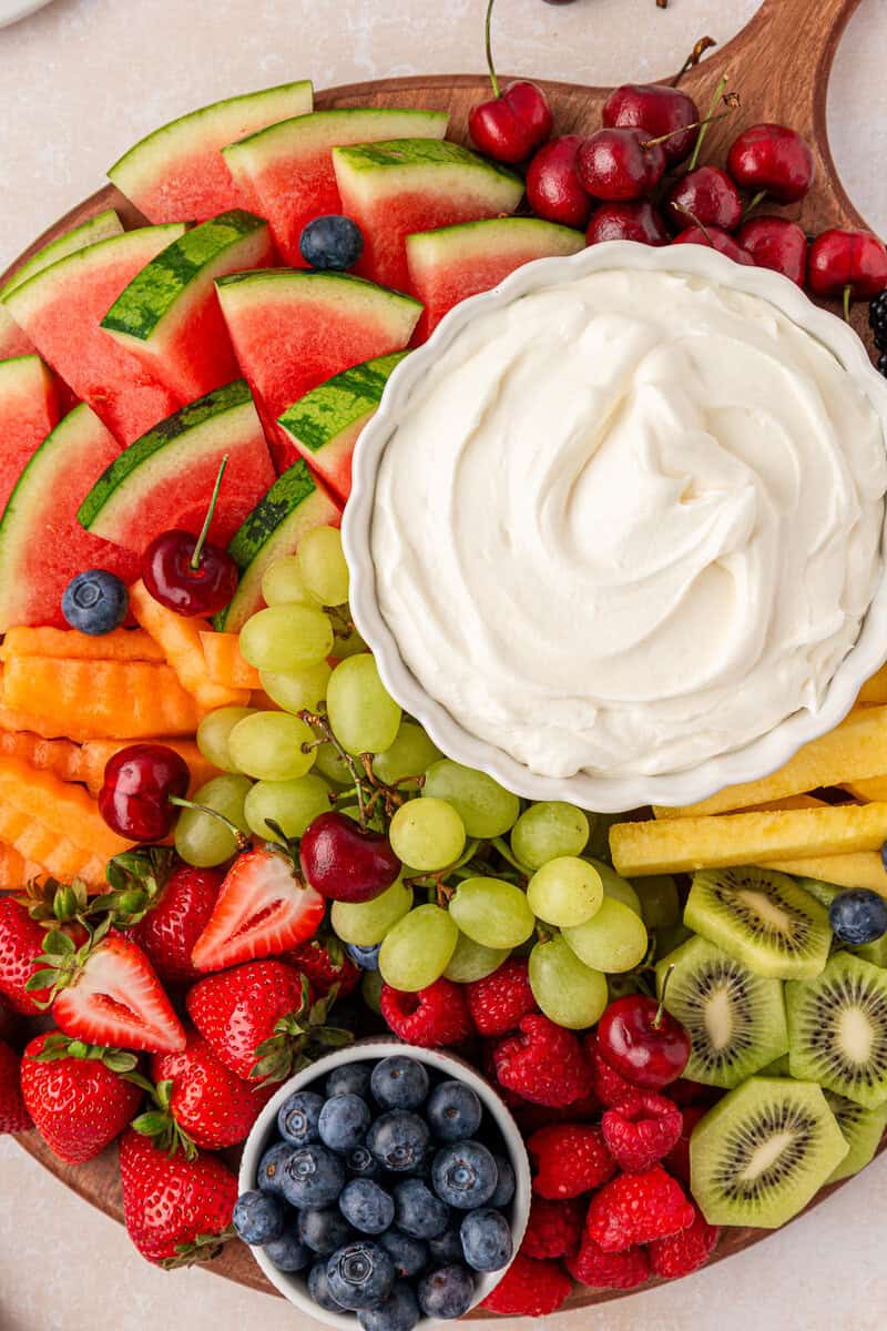 A wooden platter filled with assorted fresh fruits—watermelon, strawberries, grapes, kiwi, blueberries, raspberries, cherries, and cantaloupe—arranged around a bowl of creamy white fruit dip.