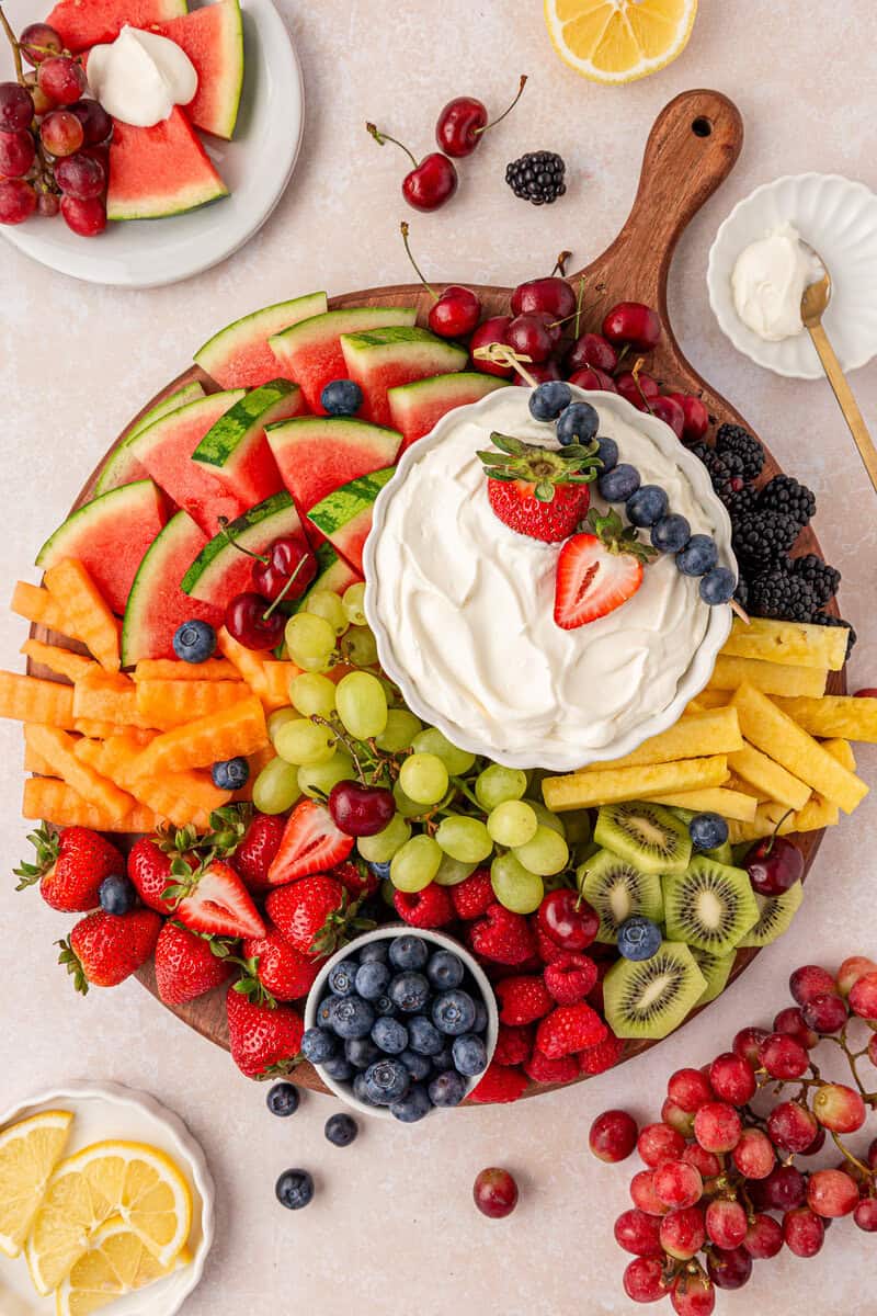 A round wooden board filled with assorted fresh fruits—strawberries, grapes, blueberries, kiwi, watermelon, cantaloupe, and mango—surrounding a bowl of creamy white dip topped with berries.