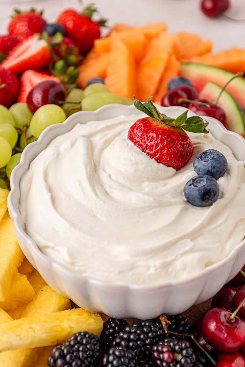 A white bowl of creamy fruit dip topped with a strawberry and two blueberries, surrounded by assorted fresh fruits including grapes, blackberries, pineapple, cantaloupe, strawberries, and cherries.