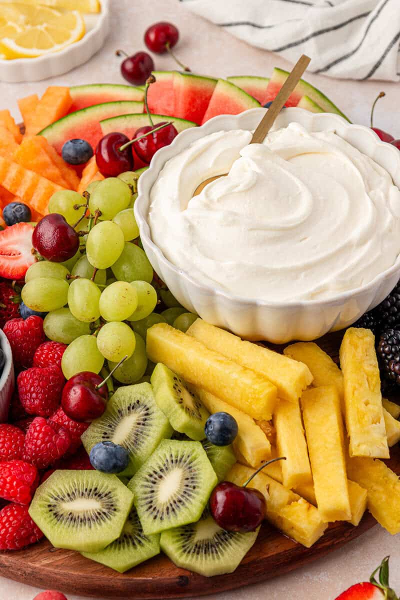 A platter of assorted fresh fruits including kiwi, pineapple, grapes, watermelon, strawberries, raspberries, blueberries, cherries, and blackberries, arranged around a bowl of creamy fruit dip with a spoon.