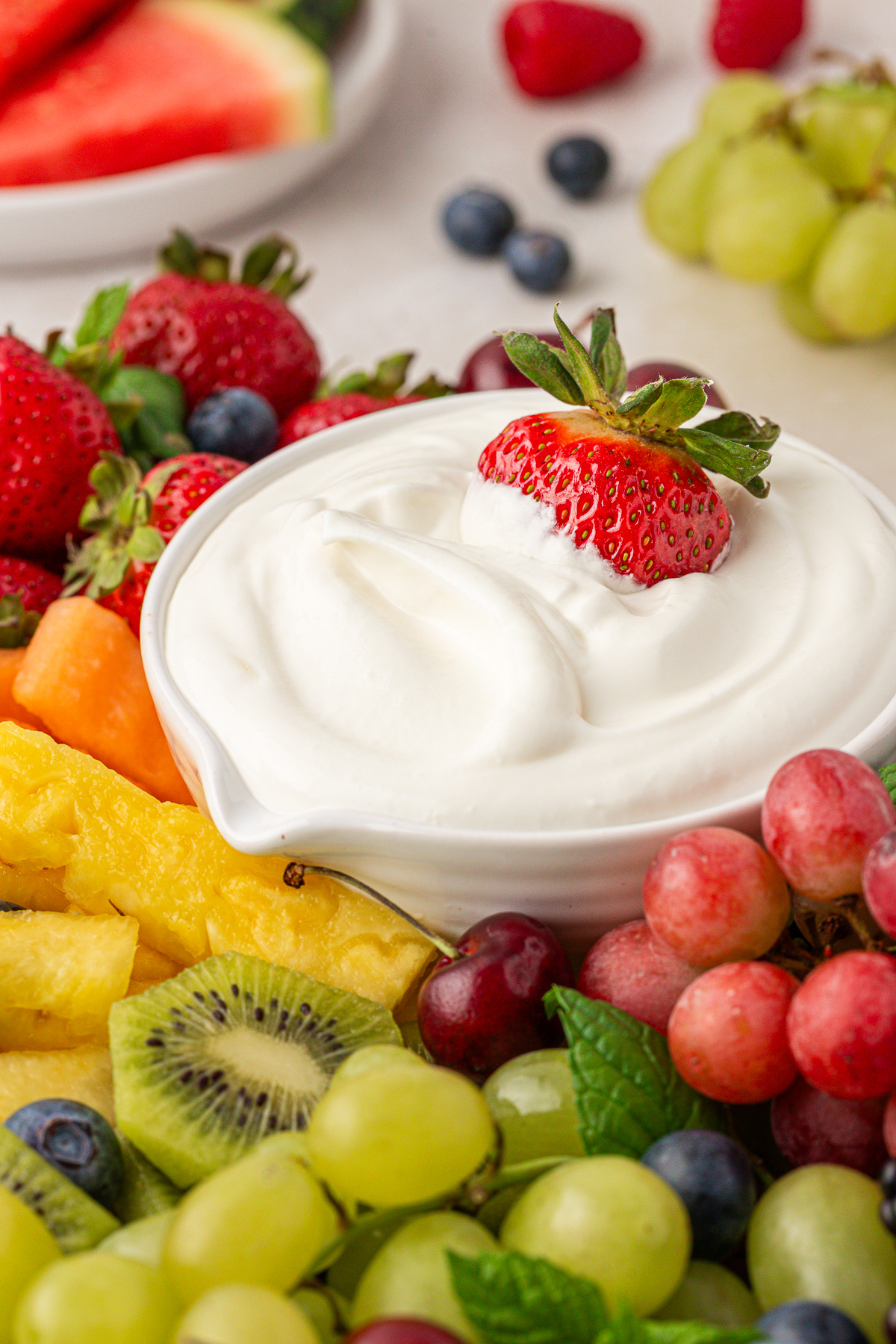 A bowl of creamy white dip is surrounded by fresh fruits, including strawberries, grapes, kiwi, pineapple, and blueberries. One strawberry is dipped in the bowl, and watermelon slices are visible in the background.