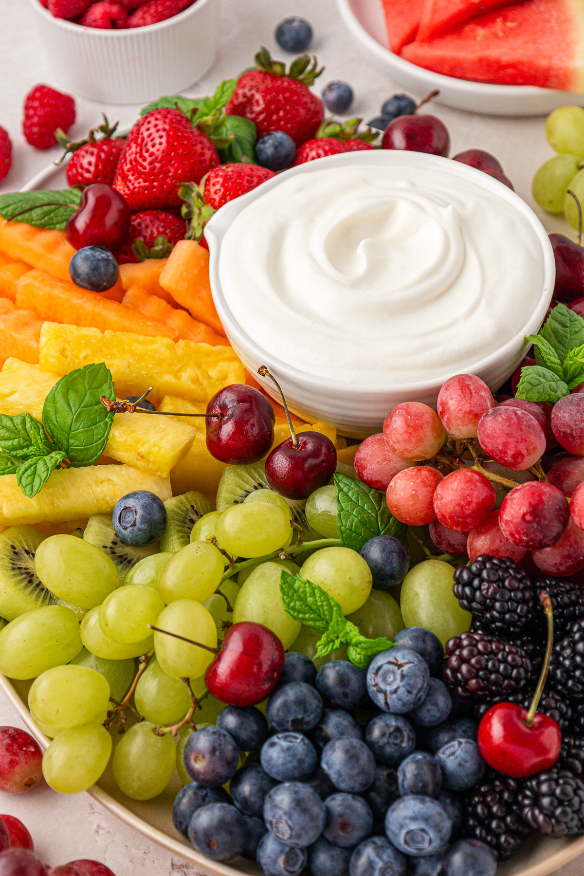 A platter of fresh fruit, including grapes, blueberries, blackberries, cherries, strawberries, pineapple, and melon slices, surrounds a bowl of creamy white dip. Mint leaves are used as garnish.
