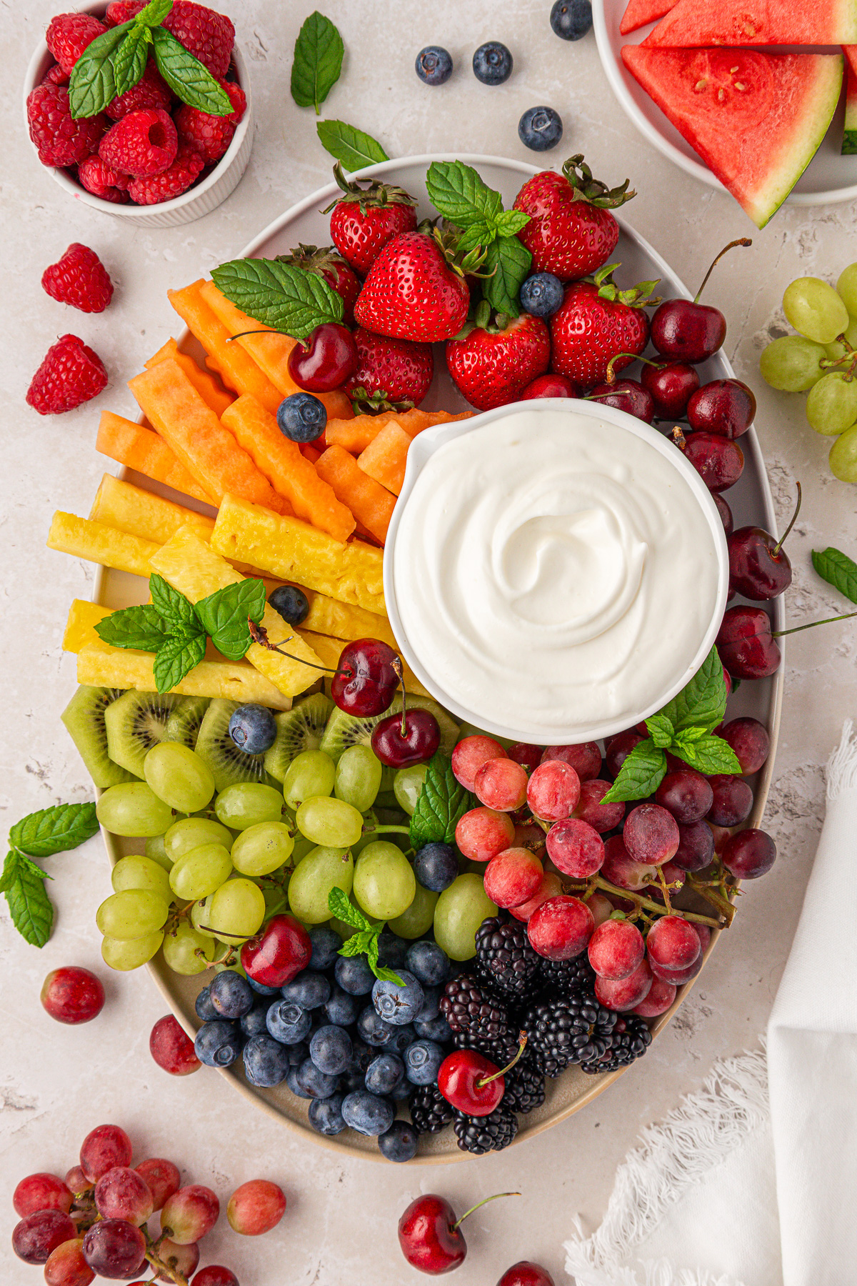 A platter with assorted fresh fruits—strawberries, grapes, blueberries, blackberries, watermelon, cherries, cantaloupe, and pineapple—garnished with mint leaves, and a bowl of creamy white dip in the center.