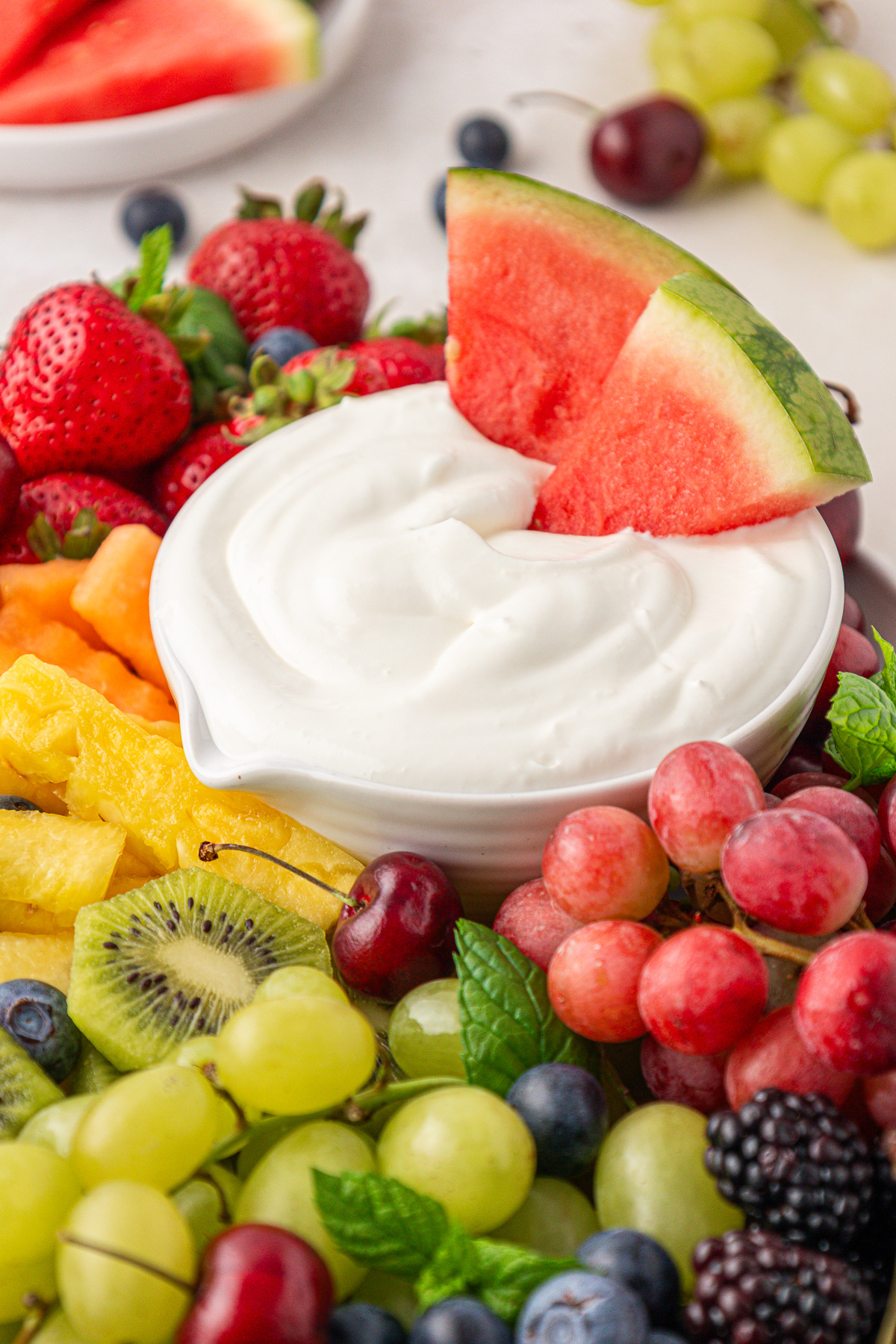 A bowl of creamy white yogurt dip garnished with watermelon slices is surrounded by assorted fresh fruits, including strawberries, grapes, blackberries, blueberries, kiwi, pineapple, cantaloupe, and cherries.
