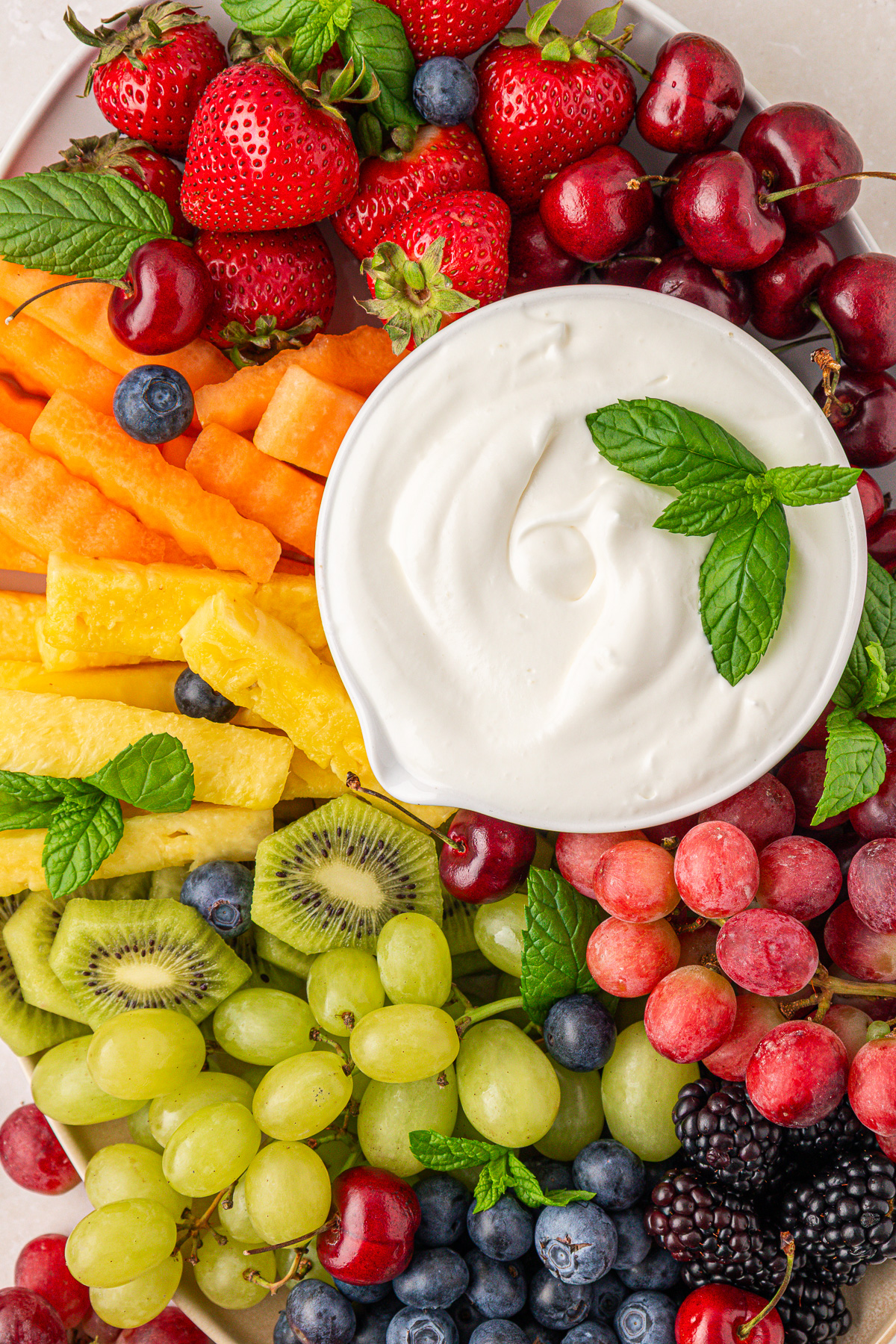 A colorful fruit platter with strawberries, cherries, cantaloupe, pineapple, kiwi, grapes, blueberries, and blackberries, arranged around a bowl of creamy white dip garnished with fresh mint leaves.