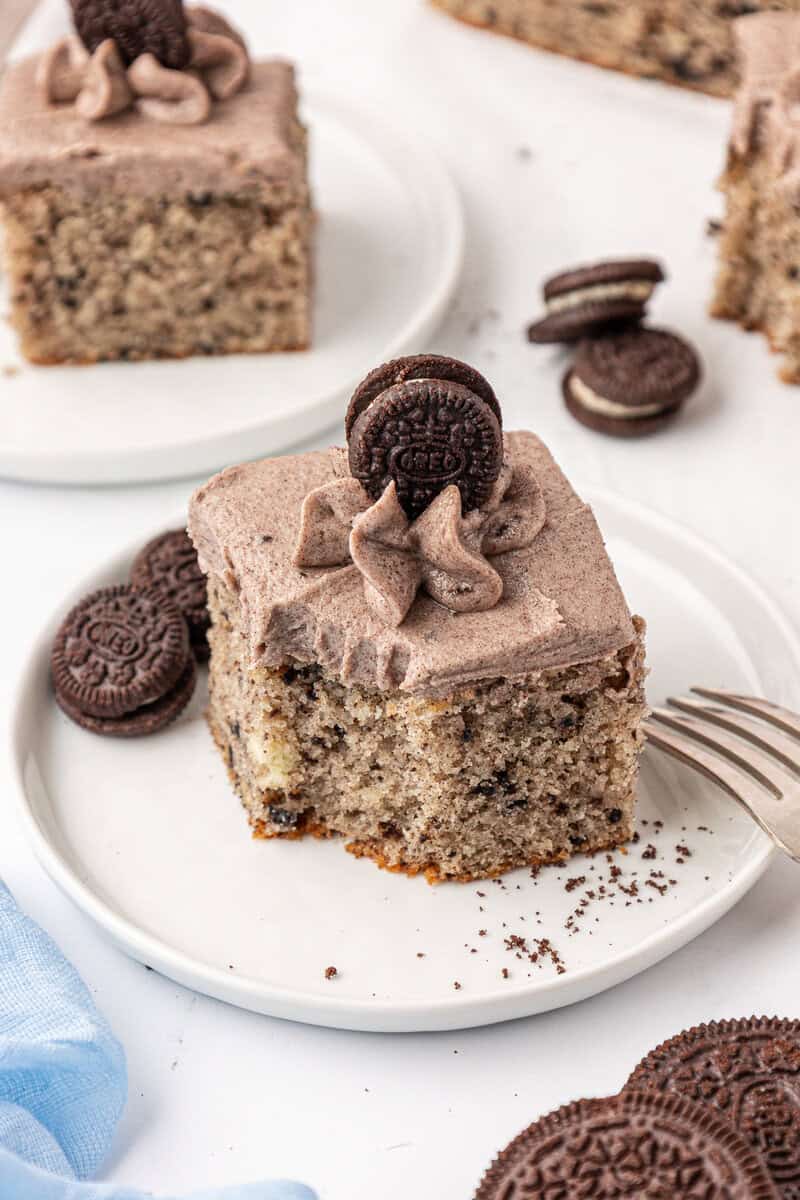 A slice of cookies and cream cake with cookies and cream frosting, topped with a mini chocolate sandwich cookie, sits on a white plate with additional mini cookies beside it and a fork nearby.