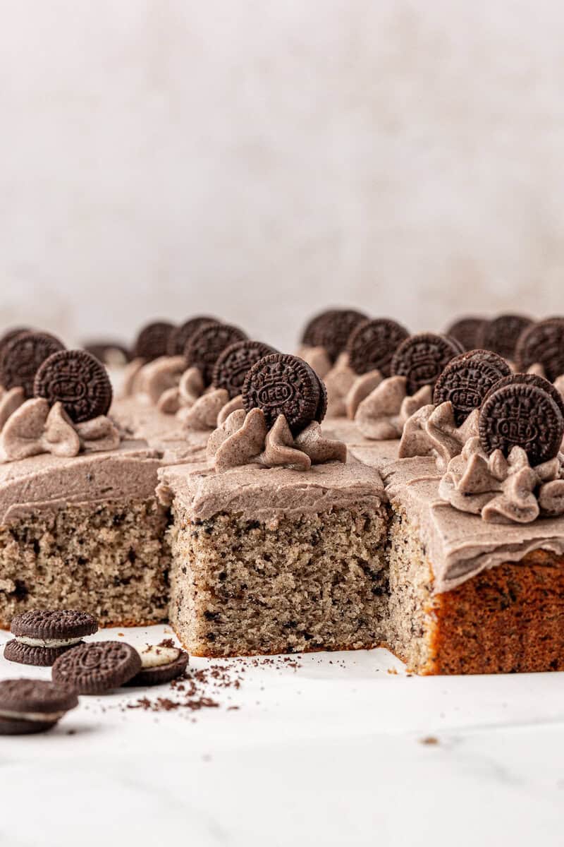 A close-up of a frosted cake topped with swirls of cookies and cream frosting, each swirl garnished with a mini chocolate sandwich cookie. A few cake slices are cut and some mini cookies are scattered nearby.