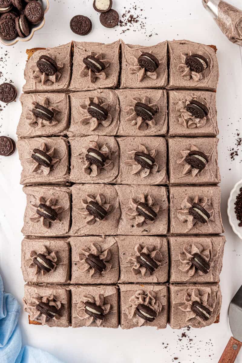 A top-down view of 20 square dessert bars with cookies and cream frosting, each topped with a swirl of frosting and a mini chocolate sandwich cookie. Crumbs and cookies are scattered around on a white surface.
