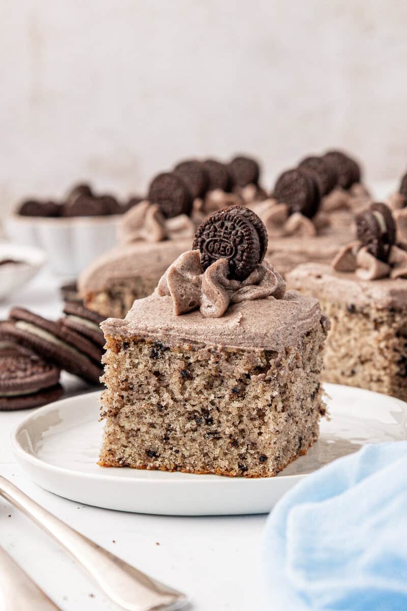 A square slice of cookies and cream cake topped with cookies and cream frosting and a mini chocolate sandwich cookie, served on a white plate with more cookies and cake in the background.