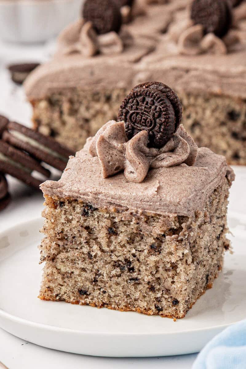 A square slice of cookies and cream cake with speckled frosting and a mini chocolate sandwich cookie on top, served on a white plate. The cake is moist with visible cookie pieces throughout.