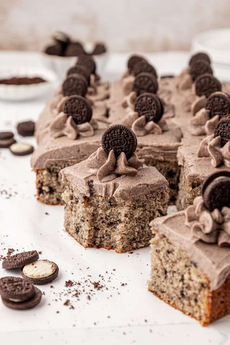 Squares of cookies and cream cake topped with cookies and cream frosting and mini chocolate sandwich cookies, arranged on a white surface with cookie crumbs scattered around.