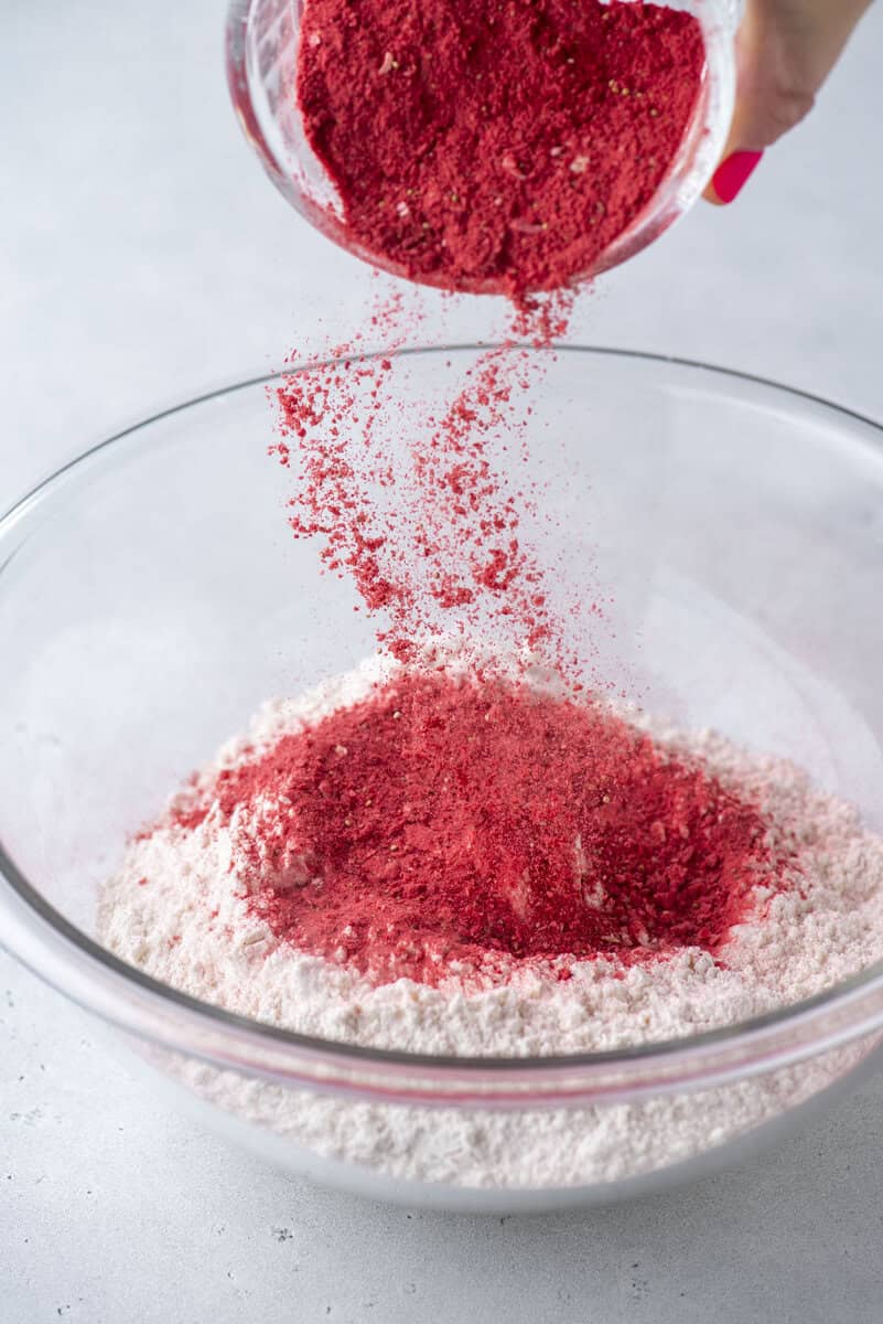 A hand pours bright red powder into a glass bowl filled with white flour, creating a colorful contrast on a light background.