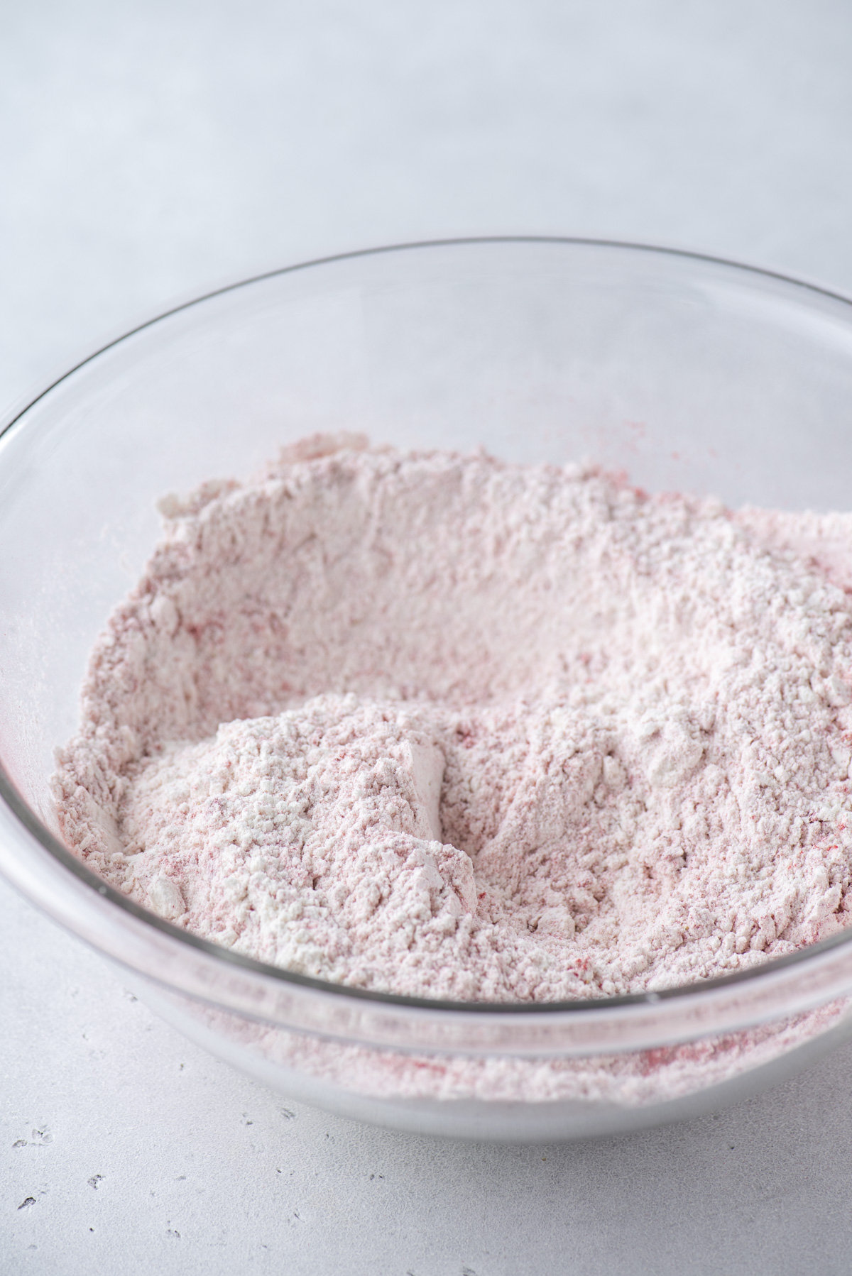 A clear glass mixing bowl contains a pale pink powdery mixture, likely baking ingredients such as flour blended with something pink, set on a light-colored surface.