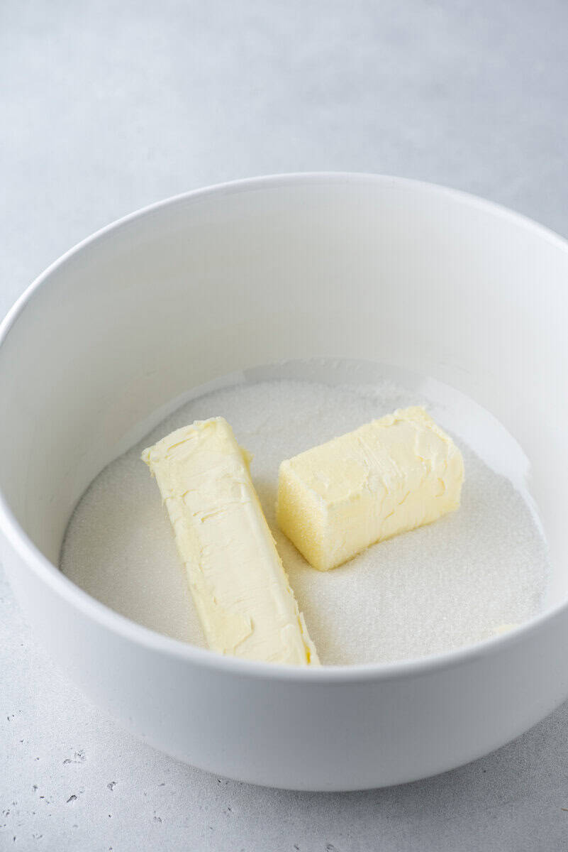 Two sticks of butter and a layer of granulated sugar are placed in a large white mixing bowl on a light gray surface.