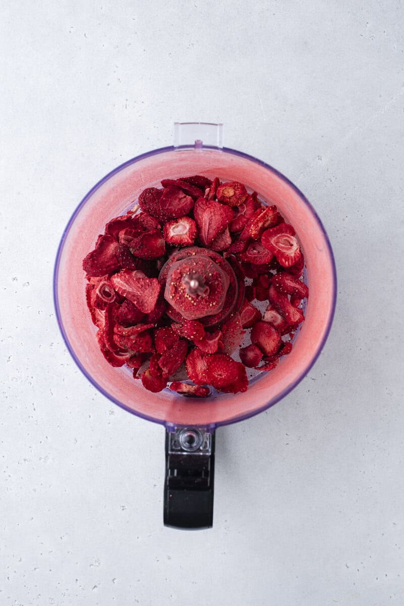 A food processor bowl filled with freeze-dried strawberries, viewed from above, on a light gray countertop.