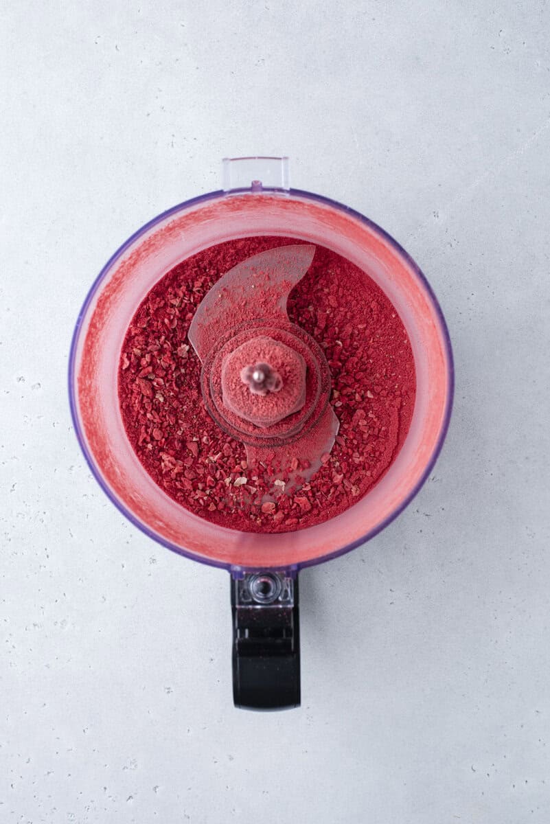 A food processor viewed from above, containing finely ground red powder, likely crushed freeze-dried strawberries, against a light gray background.